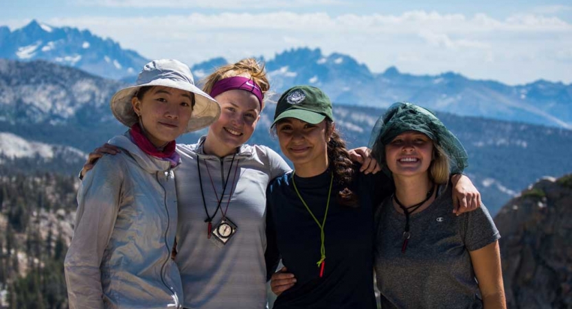 Four people stand with their arms around each other. There is a mountain landscape in the background. 